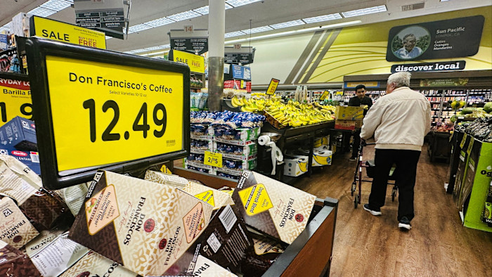 Coffee for sale is priced at $12.49 in a supermarket, with shoppers and banana displays visible in the background.