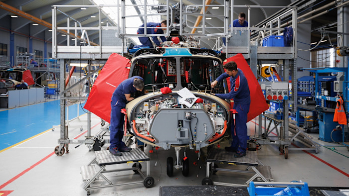 Workers assemble parts and wiring inside the nose of a Leonardo AW139 helicopter on the factory floor, using tools and reference documents.
