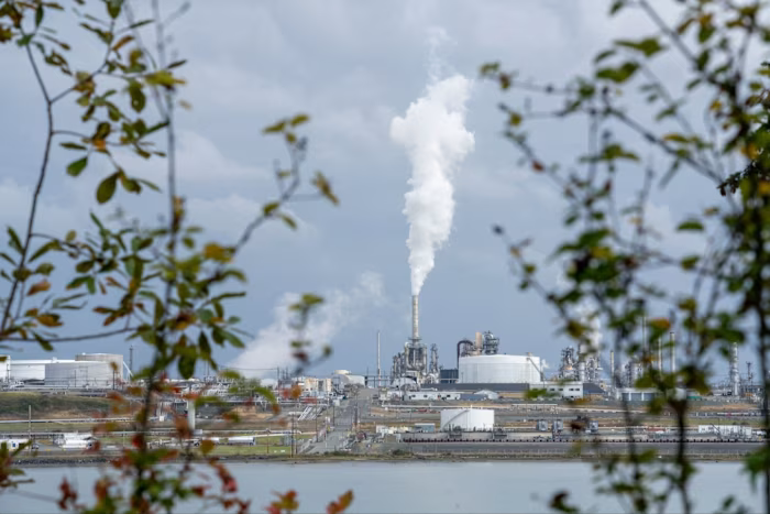 Steam or smoke rises from a stack at a US refinery, seen through branches across the water.